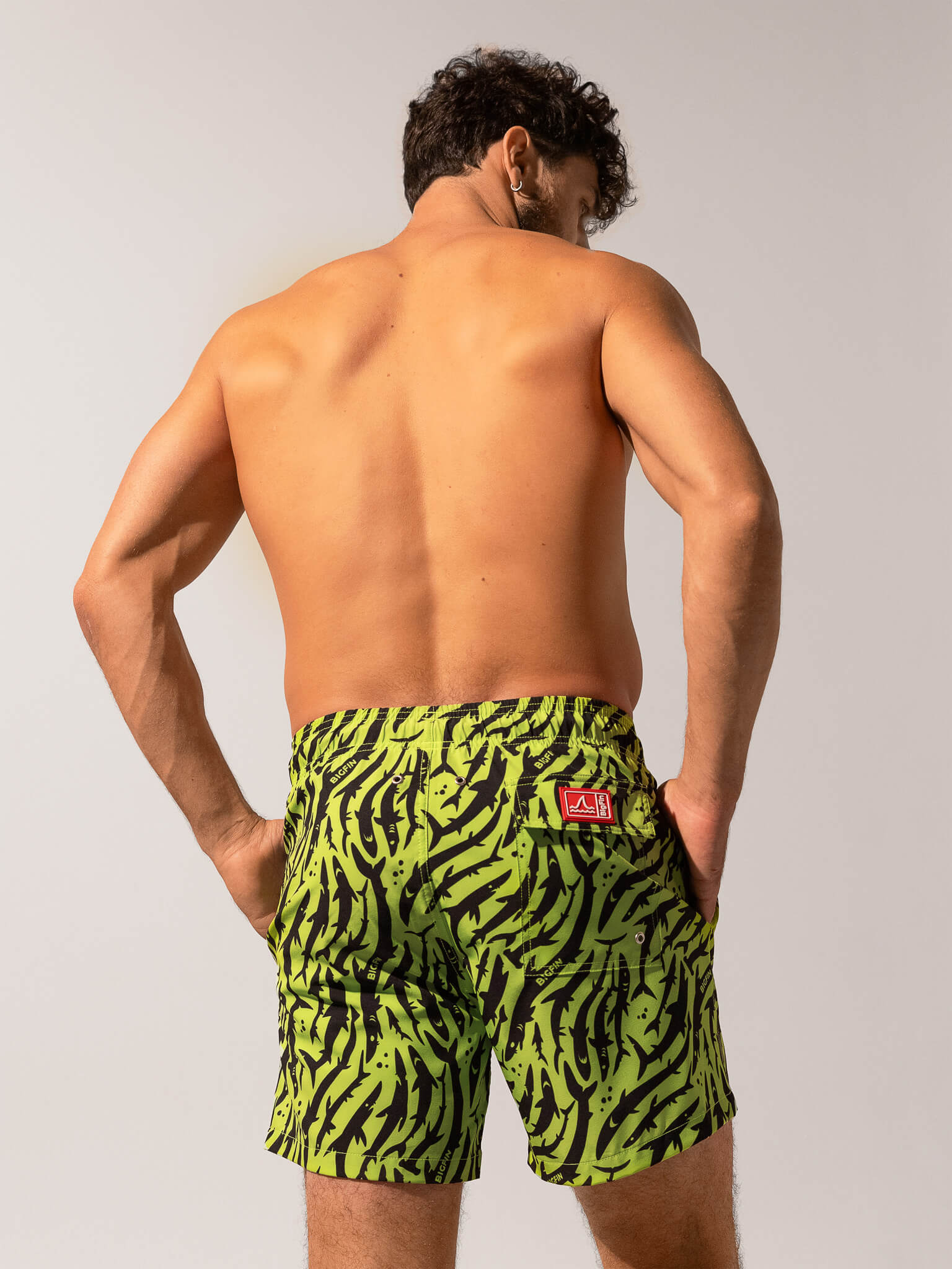 Man wearing green and black patterned swim shorts with a visible brand logo on a plain background